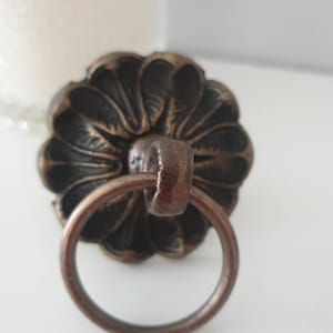 以下が含まれることがあります： 花柄のデザインとリングハンドルが付いた、ブロンズ色の装飾的なドアノブです。