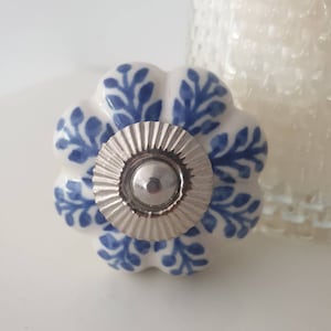 Könnte beinhalten: Weißer Keramik-Knopf mit blauem Blumenmuster und silbernem Metallzentrum. Der Knopf hat eine abgerundete, kürbisartige Form.