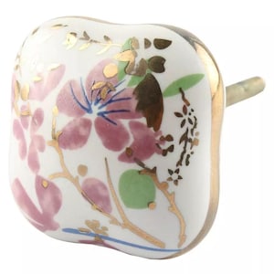 May include: A square, white ceramic cabinet knob with a floral design. The knob features pink and gold floral accents, with a gold rim and a metal stem for attachment. The design includes pink flowers, green leaves, and gold branches.