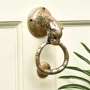 May include: A brass door knocker with a tiger head design. The knocker has a circular handle and is mounted on a round plate. The tiger's head is detailed with fur and facial features. A decorative piece of door hardware.