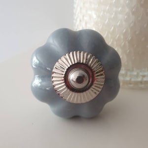 May include: A gray ceramic cabinet knob with a silver metal center. The knob has a scalloped edge and a raised, circular design in the center.