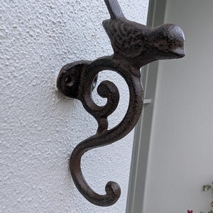 May include: A dark brown cast iron wall hook featuring a bird design. The hook is mounted on a textured white wall. The bird is perched on a decorative, swirling branch. The hook is made of cast iron.