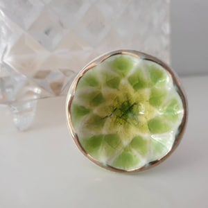 May include: A green and white ceramic knob with a gold rim. The knob has a floral design with a raised center.