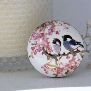 May include: A white ceramic knob with a detailed illustration of two birds on a branch with pink blossoms. The knob has a gold rim. The background is soft and blurred. The knob is likely for a cabinet or drawer.