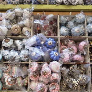 May include: Assortment of ceramic cabinet knobs in various colours and patterns, including blue, pink, white, and red. The knobs are displayed in a wooden box with multiple compartments.