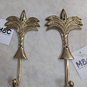 May include: Two brass-coloured palm tree wall hooks. Each hook features a detailed palm frond design at the top, a conical base, and a curved hook at the bottom. The hooks are designed for hanging items and add a decorative touch.
