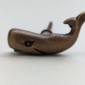 May include: Bronze-colored whale-shaped cabinet knob. The knob features a detailed design of a whale with a tail fin and a small eye. The knob is made of metal and has a vintage or antique appearance. The knob is designed to be attached to a cabinet or drawer.