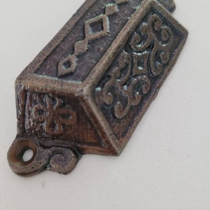 May include: A cast iron drawer pull with an ornate design. The pull is a dark brown color with a distressed finish. It has a diamond pattern on the top and a floral design on the sides.