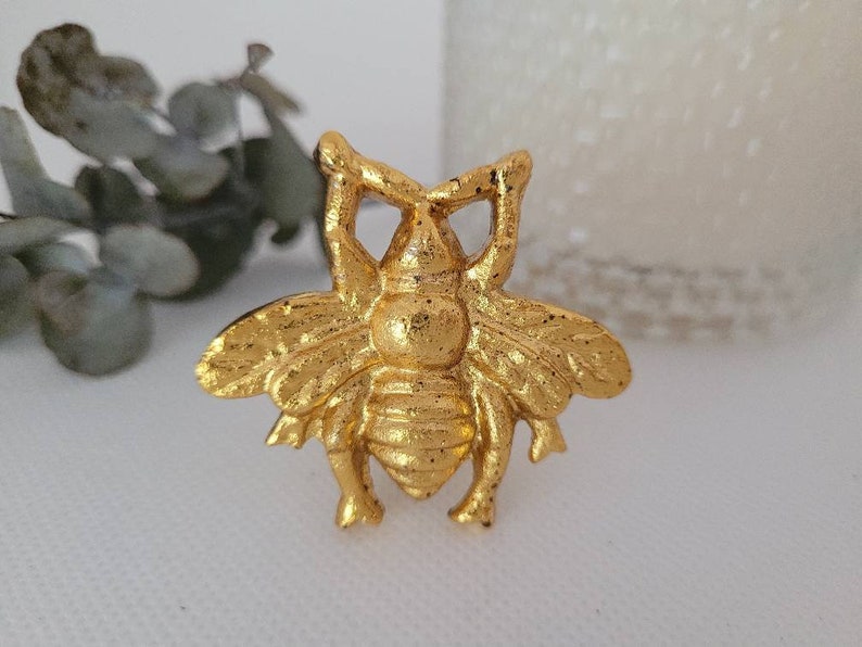 May include: A gold bee-shaped decorative object with intricate details. The bee has a raised body and wings, and its eyes are prominent.