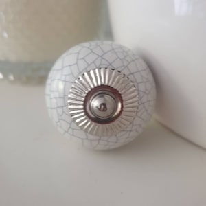 May include: White ceramic cabinet knob with a silver, decorative, circular design. The knob has a crackled finish.