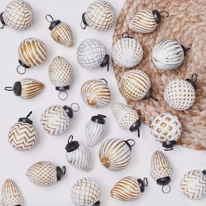 May include: A selection of Christmas baubles in various shapes, including spheres and teardrops. The baubles are white with gold or silver accents and black metal caps. Some have striped, dotted, or geometric patterns. The baubles are arranged on a white surface.