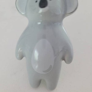 Puede incluir: Una figurita de koala de cerámica gris con ojos negros y vientre blanco. La figurita está de pie con los brazos a los lados.