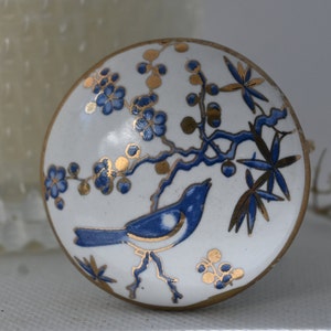 Könnte beinhalten: Ein weißer Keramik-Knopf mit einem blauen und goldenen Blumendesign, das einen blauen Vogel zeigt. Der Knopf hat einen goldenen Rand.