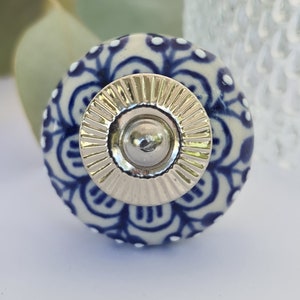 May include: A white ceramic knob with a blue and white floral pattern and a silver metal centre. The knob is round and has a raised edge.