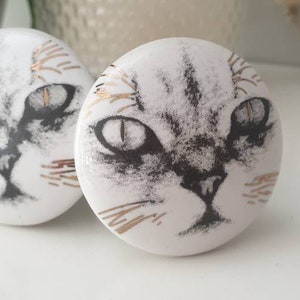 May include: Two white ceramic knobs with a black and white cat face design. The cat's eyes are a light gray color with gold accents around the eyes and whiskers.