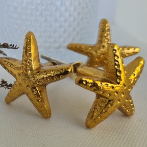 Może przedstawiać: Dwa złote, gwiazdkowate gałki do szafek. Gałki mają fakturowaną powierzchnię i są gotowe, aby dodać marynistyczny akcent do wystroju Twojego domu.