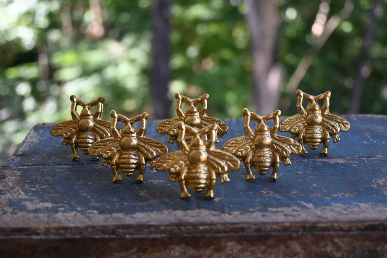May include: Six gold bee-shaped drawer pulls. The pulls are arranged in a row on a wooden surface.
