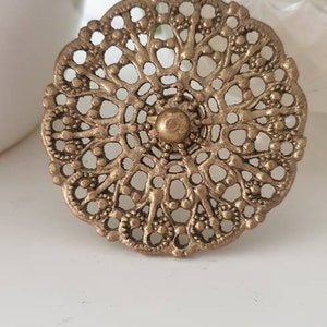 May include: A gold-toned metal decorative knob with a circular, intricate, filigree design. The knob has a raised centre with a small, round, gold-toned button.