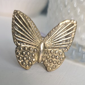 Può includere: Pomo per armadio a forma di farfalla dorata con superficie testurizzata. Le ali sono dettagliate con un motivo a ventaglio e il corpo è ricoperto da piccoli cerchi.