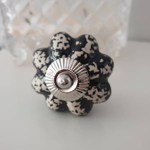 Könnte beinhalten: Schwarzer und weißer Keramik-Schrankknopf mit einem silbernen Metallzentrum. Der Knopf hat einen gewellten Rand und ein gesprenkeltes Design.