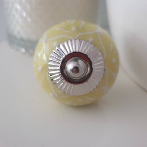 May include: A yellow ceramic knob with a white floral pattern and a silver metal centre.