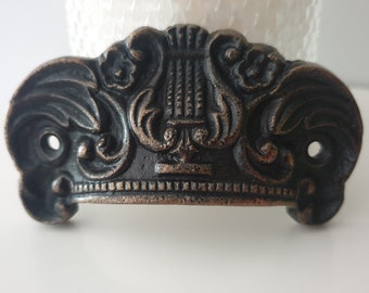 May include: A black and bronze metal drawer pull with an ornate design featuring a lyre. The pull has a rounded shape and a decorative border.