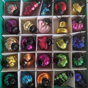May include: A collection of vintage Christmas ornaments in a divided box. The ornaments are various shapes and colors, including red, blue, gold, and purple. Each ornament has a black metal cap and loop for hanging. The box is divided into individual compartments.