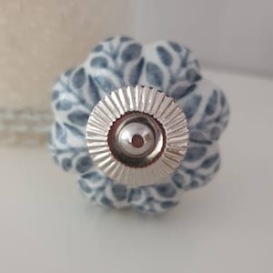 Könnte beinhalten: Ein weißer Keramik-Knopf mit einem blauen Blumenmuster und einem silbernen Metallsockel. Der Knopf hat eine erhabene, strukturierte Oberfläche.