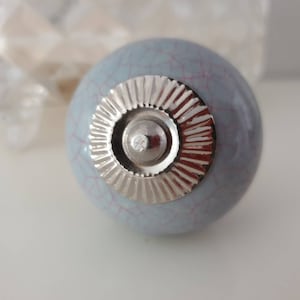 Peut inclure: Un bouton en céramique bleu clair avec un centre en métal argenté. Le bouton a une finition craquelée et un design décoratif.
