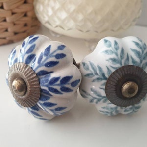 以下が含まれることがあります： 白いベースと花柄のデザインが施されたセラミック製のキャビネットノブ2個。1つのノブは青い花柄のアクセント、もう1つのノブは緑の花柄のアクセントが付いています。両方のノブは真鍮色のセンターが付いています。