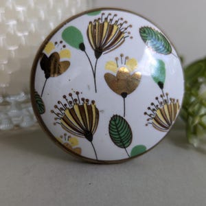 May include: A white ceramic knob with a gold rim and a floral design featuring yellow, green, and brown flowers.