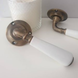 Op de afbeelding: Twee antieke messing deurgrepen met witte keramische knoppen. De grepen hebben een traditioneel ontwerp en zijn perfect om elke kamer een vleugje elegantie te geven.