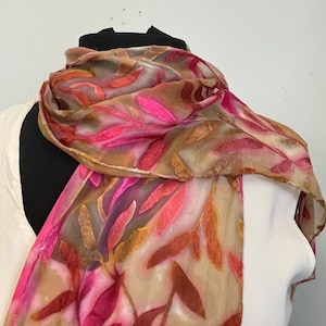 May include: A sheer scarf with a leaf pattern in shades of pink, orange, and brown. The scarf is draped over a mannequin, showcasing its flowing fabric and vibrant colors. The design features a mix of warm and cool tones.