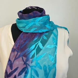 May include: A sheer scarf in shades of blue and purple, draped over a mannequin. The scarf features a leaf pattern in a lighter shade of blue, creating a contrast against the darker background. The scarf is a fashion accessory.