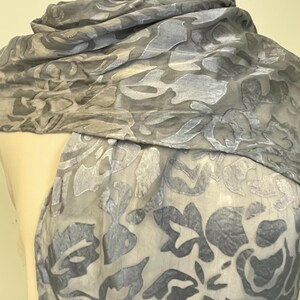 May include: A sheer, gray scarf with a floral pattern. The scarf is draped over a mannequin, showcasing its flowing fabric and design. The floral pattern is a darker shade of gray, creating a subtle contrast.