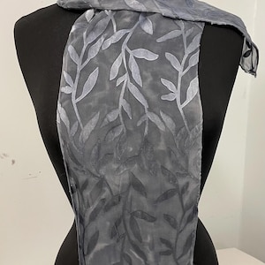May include: A grey silk scarf with a leaf pattern. The scarf is draped over a black mannequin.