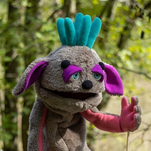 May include: A plush puppet with a gray body, purple accents, and teal hair. The puppet has a pink arm and is held by a wooden stick. The puppet's eyes are blue, and it has a black nose. The background is a blurred green forest.
