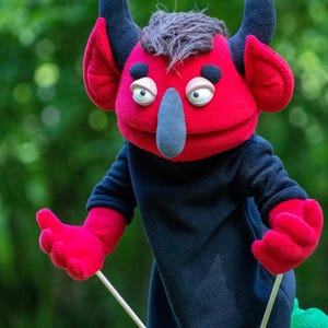 May include: A red and black fleece hand puppet featuring a creature with two dark horns, large ears, a grey nose, and grey hair. The puppet has a black body, red hands, and is controlled by two thin wooden rods attached to the arms for movement.