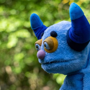 Op de afbeelding: Een blauwe pluche pop met grote, expressieve ogen en een roze neus. De pop heeft twee donkerblauwe hoorns en een geel en oranje oogontwerp. De achtergrond is een wazig groen.