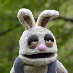 Könnte beinhalten: Eine Plüsch-Hasenpuppe mit cremefarbenem, strukturiertem Äußeren. Die Puppe hat große, schlaffe Ohren mit braunen Innenfutter, braunen und grauen Augenakzenten und eine rosa Nase. Sie trägt eine graue Latzhose.