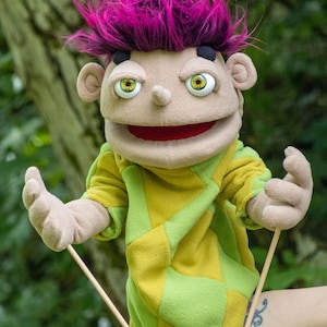 May include: A hand puppet with bright magenta spiky hair, tan skin, and large green eyes. It features a wide red mouth and wears a long-sleeved shirt with a yellow and lime green checkered pattern. Two thin wooden rods are attached to its hands.