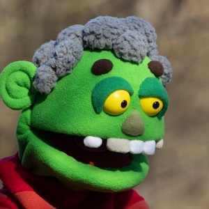 May include: A green puppet head with yellow eyes, white teeth, and grey hair. The puppet has a red and orange striped shirt. The puppet's face is green with brown spots above the eyes. The puppet has a large open mouth.