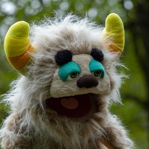 May include: A plush monster puppet with shaggy, light brown fur. It has large, yellow horns with orange accents, black and teal eye details, and a red mouth. The puppet is set against a blurred green background.