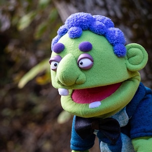 Puede incluir: Un títere verde con cabello morado texturizado, cejas moradas y ojos blancos grandes. El títere presenta una boca ancha con dos dientes blancos visibles, un interior rojo y viste una camisa azul oscuro con una pajarita negra en el cuello.