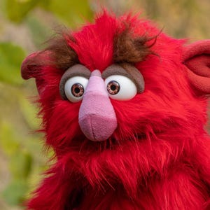 以下が含まれることがあります： 茶色の毛皮のアクセントと大きく表現力豊かな目を持つ赤い人形のクローズアップ。人形はピンクの鼻と赤い耳をしています。人形の毛皮は鮮やかな赤色で、気まぐれで遊び心のある外観を作り出しています。