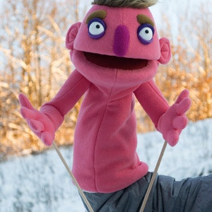 A pink  professional hand puppet with brown eyebrows, a purple nose, and large white eyes. The puppet has arm rods .