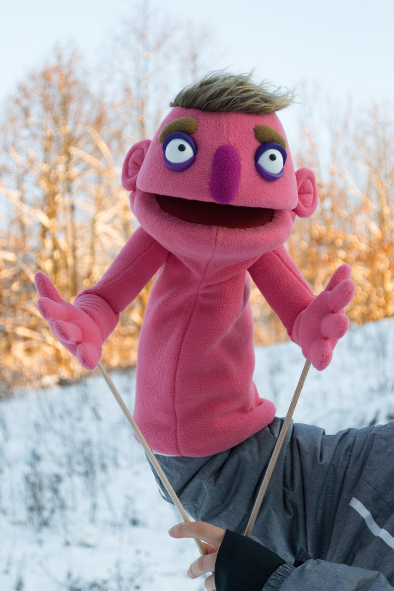 A pink  professional hand puppet with brown eyebrows, a purple nose, and large white eyes. The puppet has arm rods .