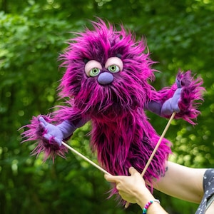 May include: A fuzzy purple monster puppet with long, shaggy fur, large pale pink eyelids, and bright green eyes. The creature features a round purple nose and purple fabric hands. Two thin wooden control rods are attached to the puppet's arms.