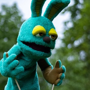 May include: A teal blue puppet with long ears, yellow eyes, and a brown nose. The puppet is holding two white sticks.