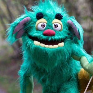 Puede incluir: Una marioneta de monstruo color verde azulado con pelaje largo y desgreñado, dos cejas de pompón negro, ojos grandes blancos con iris marrones, nariz roja y una boca ancha con dientes visibles. Tiene brazos y manos verde claro y orejas moradas.